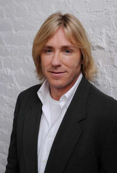 Actor, Ron Eldard giving a pose in a black coat and white shirt.