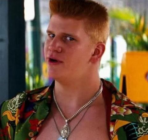 A white man with red hair and a colorful shirt with lots of neck jewelery.