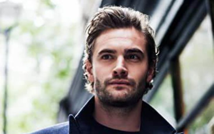 Tom Bateman wearing a black cardigan and posing for a photo.